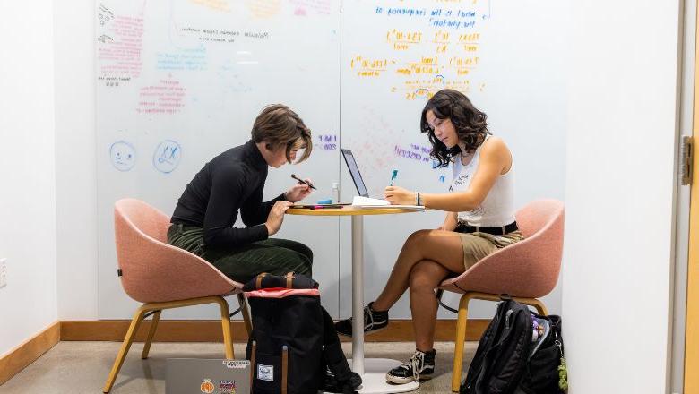 Two students sitting in front of a white board studying. 